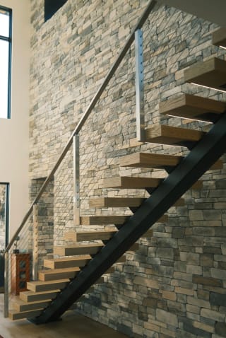 Light oak treads on a black mono stringer with stainless cable railing climb a stacked-stone wall.