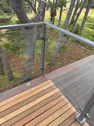 Corner of a post-to-post glass railing with gray cap rail above a hardwood deck and wooded yard.