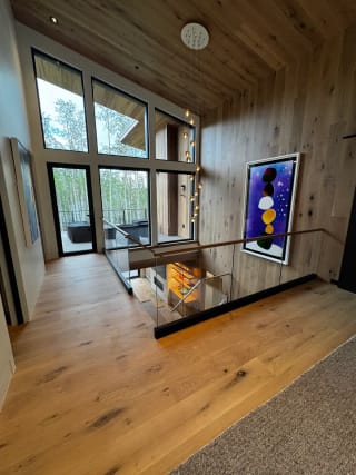 Frameless glass in a black base channel with a wood handrail surrounds a stairwell in a wood-paneled great room with tall windows.