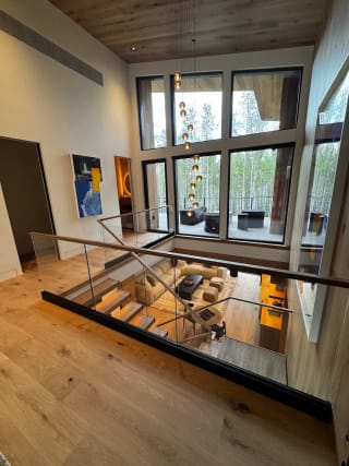 A frameless glass guard with wood cap overlooks a double-height living room with floor-to-ceiling windows.