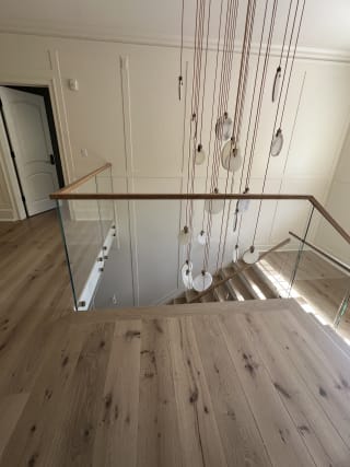 A frameless glass guard with an oak cap wraps a stair opening beneath a cascading pendant light in a paneled hallway.