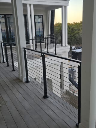 Black aluminum posts with stainless cable infill run along a covered wood-deck porch with white columns.