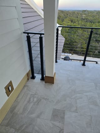 Black-post cable railing edges a stone-tile covered balcony beside white columns and a treetop view.