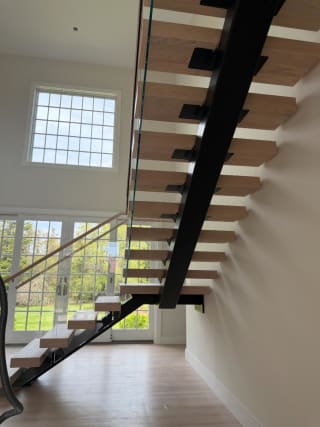 View under a black mono stringer carrying white-oak treads with glass railing in front of tall grid windows.