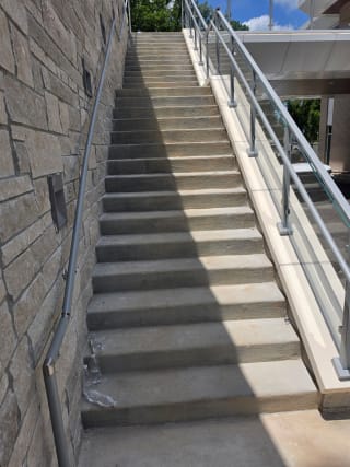 Post-to-post glass railing with stainless handrail runs up a long exterior stone stair beside a stacked stone wall.