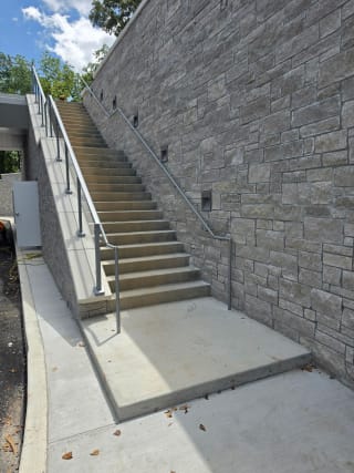A long exterior concrete stair with post-to-post glass railing and a wall handrail beside a stone retaining wall.