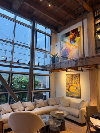 Frameless glass railing lines a timber-beamed mezzanine above a living room with tall steel windows.