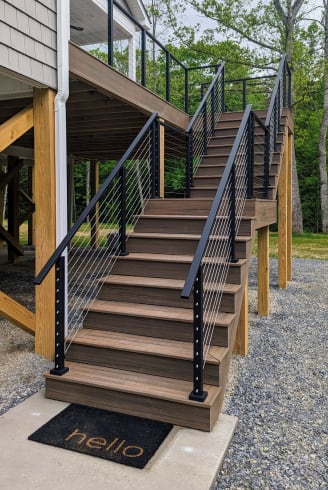 Black cable railing lines a tall composite deck staircase rising to an elevated porch over gravel.