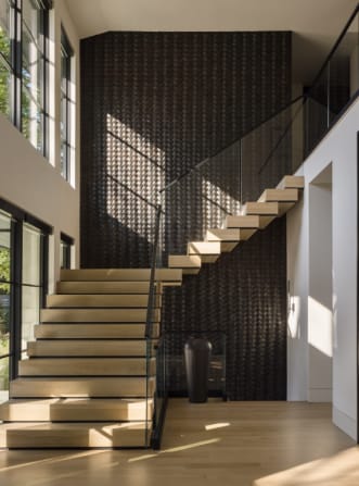 Light oak cantilevered treads with frameless glass rise along a dark woven-texture wall in a sunlit two-story foyer.