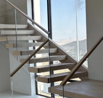 Light oak floating treads turn at a landing with frameless glass and wood handrails beside a tall window.