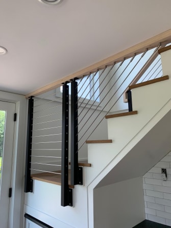 Black posts and a wood rail with stainless cables that run up into the ceiling over a white stair.
