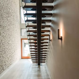 Looking up beneath a black mono stringer with oak treads between a stacked stone wall and a warm sconce-lit wall.