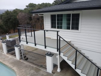 Black cable railing wraps a raised gray deck and exterior stair on a white home beside a pool.