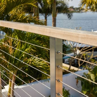 Close detail of a stainless cable post and wood cap rail overlooking palms and a waterway.
