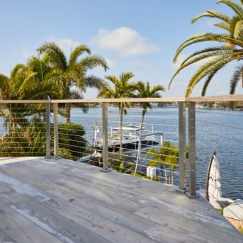 Stainless posts with cable infill and wood cap edge a weathered deck overlooking a palm-lined canal.