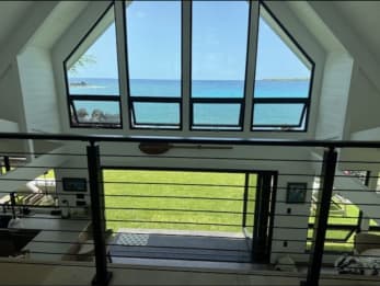 Black posts and horizontal rod infill frame a view through tall gable windows to a lawn and blue ocean.