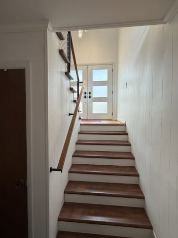 A wood handrail on black brackets runs along a white-paneled stairwell leading to a glass front door.