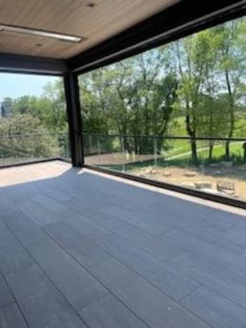 A covered porch with gray plank tile is enclosed by black-capped base-rail glass looking out to trees and a pond.