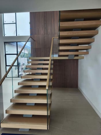 Light oak floating treads with frameless glass and a wood handrail turn at a landing against a walnut wall.