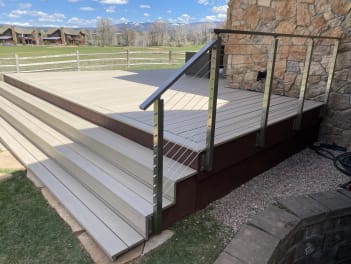 Composite deck with wide steps and stainless post cable railing against a stone chimney and mountain pasture.