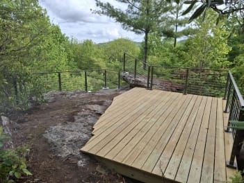 Wood platform on a rock outcrop surrounded by bronze-post cable railing overlooking forested mountains.