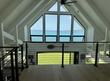 Black horizontal rod railing along an open loft under a white vaulted ceiling with a triangular wall of windows facing the ocean.