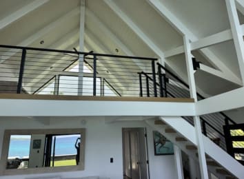Black rod railing lines a white-trimmed loft and stair beneath an exposed-beam vaulted ceiling.