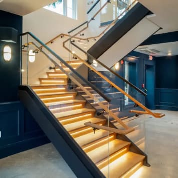 LED-lit wood treads on a dark stringer with glass railing and wood wall handrails in a navy commercial stairwell.