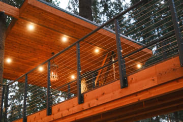 Black cable railing along a cedar treehouse deck beneath a recessed-lit wood canopy.