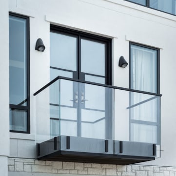 Frameless glass Juliet balcony with black top cap and fascia clamps outside double doors on a white facade.