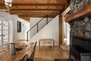 Black rod railing along a white shiplap stair beyond a rustic dining room with exposed beams and a stone fireplace.