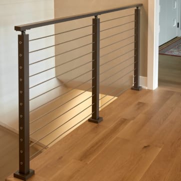 Dark bronze posts with cable infill and a matching top rail guard a stair opening on white oak floors.