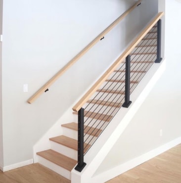 Black posts with cable infill and a light oak cap rail on a white knee wall, matched by a wall-mounted handrail.