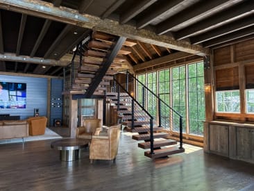 Wood floating treads on a black mono stringer switch back beneath exposed barn beams, with black cable railing and walls of windows.