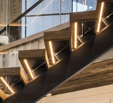 Close view of wood floating treads with vertical LED cutouts on a black stringer, with a thin glass railing edge.