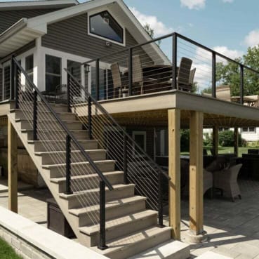 Black cable railing follows a composite staircase up to a covered elevated deck above a stone patio.