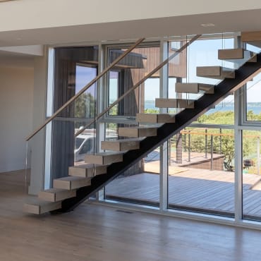 Black mono-stringer stair with thick oak treads and frameless glass rises in front of floor-to-ceiling windows.