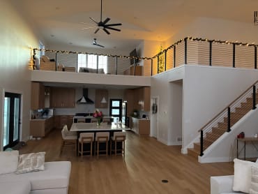 Black cable railing with a wood cap follows a stair and wraps a loft above an open kitchen and living room.