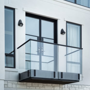A small balcony with frameless glass on black talon clamps and a black top cap below French doors on a white facade.
