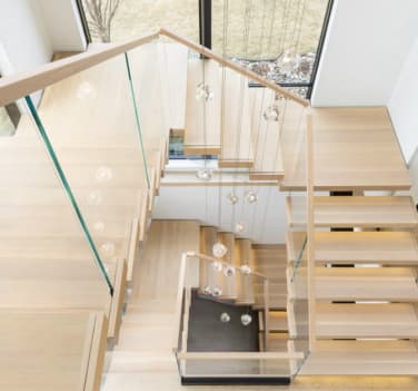 Overhead view of light oak cantilever treads turning at landings with frameless glass and oak handrails under glass pendant lights.