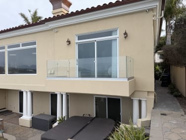 A frameless glass balcony railing on a stucco Mediterranean-style home above a stone patio.