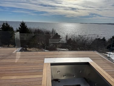 Frameless glass railing around a hardwood rooftop deck with hot tub overlooking the ocean at sunset.