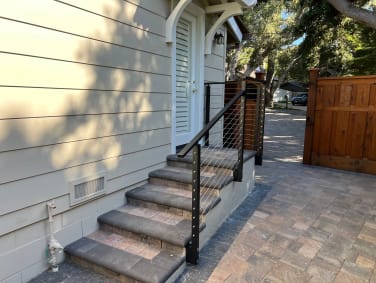 Black-post cable railing follows stone entry steps beside a white door and a cedar gate on a paver courtyard.