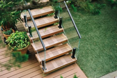 Overhead view of slatted wood treads on an exterior FLIGHT stair with black-post glass railing over a composite deck and lawn.