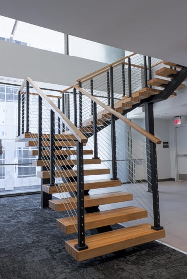 Thick oak floating treads on a black FLIGHT stringer switch back at a landing with black cable railing and oak handrails in a glass-walled office lobby.