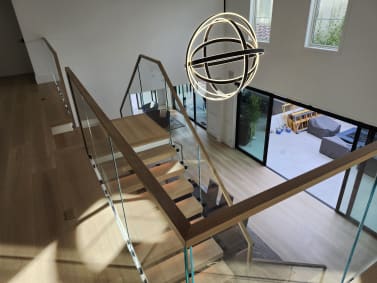Overhead view of a glass railing with a white oak handrail descending past an orb chandelier to a bright living area.