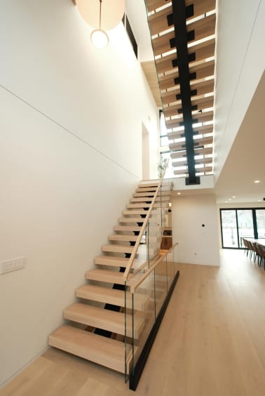 Light oak treads climb a black mono stringer through a narrow triple-height space with glass railing.