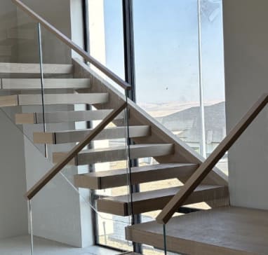 Light oak floating treads turn at a landing with frameless glass and wood handrails beside a tall window.
