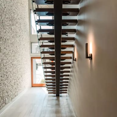 Looking up beneath a black mono stringer with oak treads between a stacked stone wall and a warm sconce-lit wall.