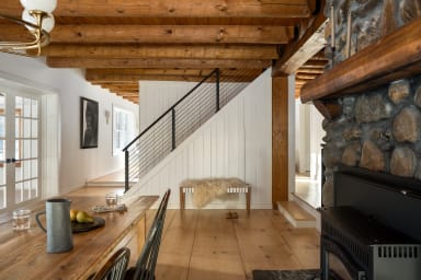 Black rod railing along a white shiplap stair beyond a rustic dining room with exposed beams and a stone fireplace.